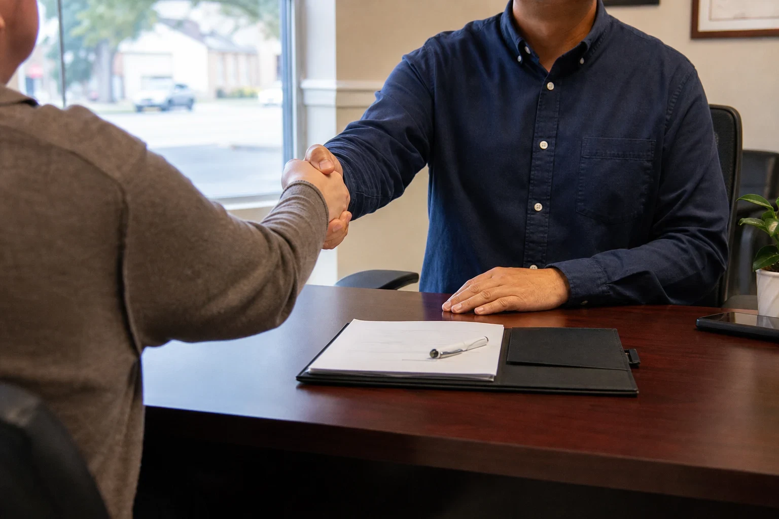 Agent in a navy button-down shirt shakes hands with a client across a wooden desk holding a folder, papers and a pen, with a street-facing window behind
